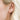 Close-up of a person with fair skin and blonde hair wearing a delicate gold ear climber earring with five small sparkling gemstones arranged in a curved line. The soft natural lighting highlights the earring’s subtle elegance, with strands of hair gently framing the ear.