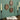 A styled setting featuring three oval wall decorations with vintage botanical illustrations, mounted on a muted green wall. Below, a dark wooden surface holds an open book with a golden cord, flanked by two tall black candlesticks with cream-colored taper candles.