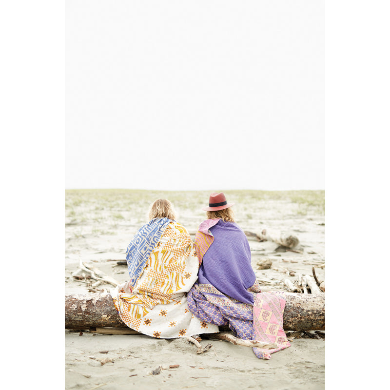 Two women sitting on a log at the beach wrapped in kantha quilts. 