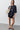 A woman modeling an oversized black t-shirt with a white teardrop graphic, paired with black shorts, black sandals, and carrying a beige shoulder bag against a light gray studio background.