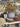 A rustic dried floral arrangement in a large gray ceramic pot, displayed on a table alongside a striped dog portrait, wooden cutting boards, braided textiles, and other home decor items in a softly blurred shop background.