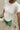 Person wearing a white t-shirt and green pants, holding a drink on sandy ground.