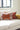 A decorative throw pillow featuring a large orange tiger print on a light pink background, styled on a white sofa next to other patterned cushions in a bright room.