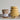 A set of four stacked measuring cups in a textured pale yellow glaze with green leaf accents, displayed next to a glass jar of Orange Marmalade and two gold spoons on a light gray surface.