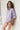 A woman leans against a white wall wearing an oversized light purple t-shirt, striped knit sleeves, and beige shorts.