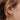 Close-up of a woman’s ear adorned with a gold floral stud earring and a textured gold ear cuff. Her light brown hair is loosely styled, with soft strands framing the jewelry. The intricate detailing of the earrings catches the light, complementing the warm tones of her hair and skin.