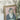 Close up of the framed vintage canvas portrait of a woman with hand resting on her face, set in an ornate antique gold frame leaning against a wall featuring cram and black floral wallpaper. 