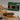 A packaged food product box for Saltspring Kitchen Co. Triple Pepper Jelly sits behind a prepared sandwich on a wooden board with potato chips, pickles, bread, and a small bowl of dark jelly on a light tabletop.