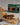 A packaged food product box for Saltspring Kitchen Co. Triple Pepper Jelly sits behind a prepared sandwich on a wooden board with potato chips, pickles, bread, and a small bowl of dark jelly on a light tabletop.