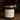 Smoke and Incense soy wax candle in an amber glass jar with gold metal lid sitting on a dark wood plank floor inside a rustic log cabin, with a wood burning stove in background. 