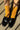 A close-up of a person's feet wearing bright mustard yellow crew socks and shiny black penny loafers, standing on a concrete surface with diagonal shadows.