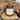 An amber glass jar candle with a label that reads 'Tonka + Tobacco', placed on a wooden surface with woven trivets in the background.