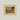 Vintage-style landscape print of a small village nestled between cliffs, in a muted earthy palette, framed in gold.