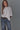 A woman wearing an oversized white short-sleeve t-shirt tucked into blue jeans with a black belt, standing against a solid gray studio background.