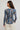 A woman shown from the back wearing a long-sleeve floral top with a blue base and multicolored floral print, paired with light beige pants, against a plain light gray studio background.
