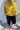 A person wearing a chunky knit mustard yellow mock neck sweater, dark navy cropped pants, and red pointed flats, standing against a white textured wall.