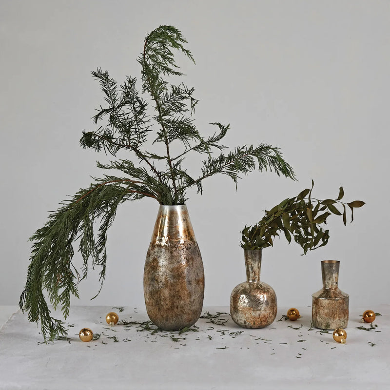 Three metal vases with distressed pewter finish styled with green branches and gold ornaments on table.  