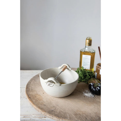 Wooden handled whisk resting on a stoneware bowl. Styled with fresh herbs and olive oil.