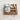 A soft, pastel green dinosaur plush toy resting inside a ribbed white fabric basket, placed on a rustic wooden tray with a second compartmentalized tray featuring colorful paint stains, set against a textured, beige backdrop.