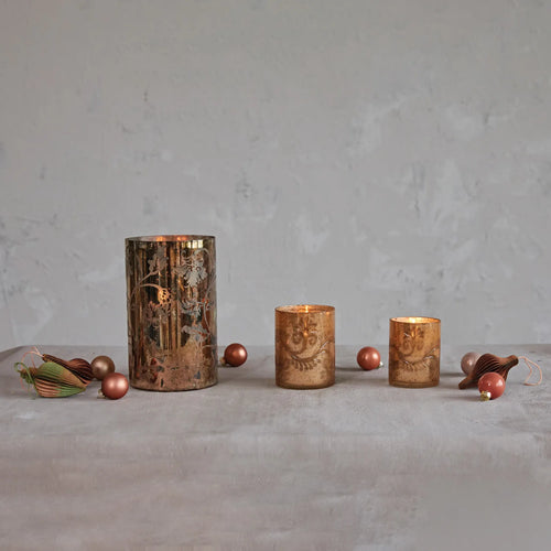 Set of 3 mercury glass votive holders glowing with tealights inside on a table with Christmas ornaments. 