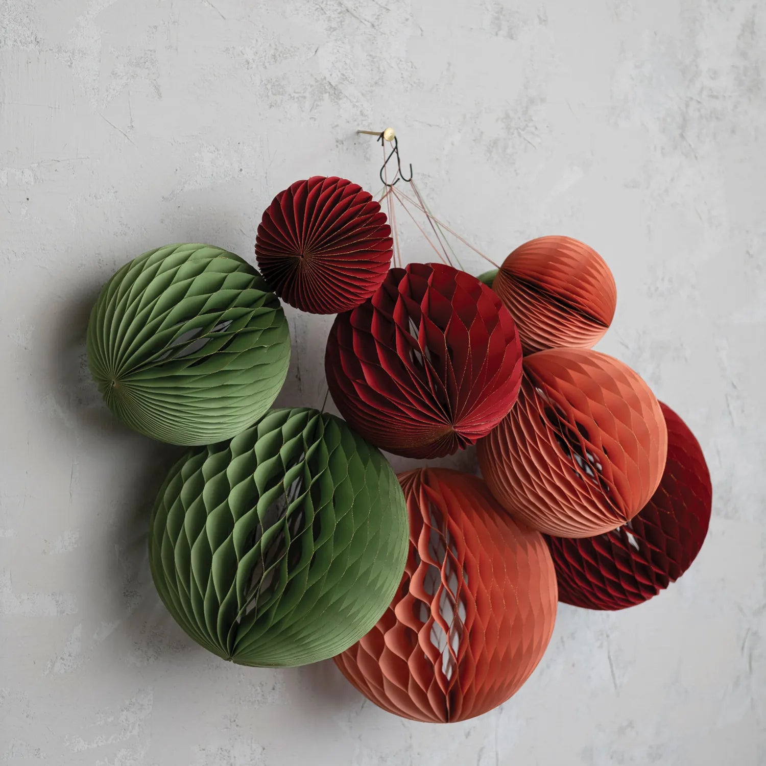 Handmade recycled paper ornaments in red, green and pink with gold edges are hung together agaist a wall.