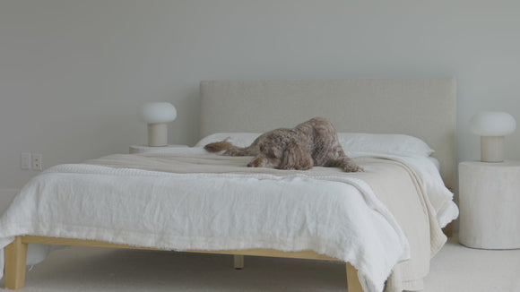 Video showcasing the natural oak platform bed with a light beige upholstered headboard, styled with white and cream bedding, a sculptural stone nightstand, and a mushroom-style table lamp in a serene, modern bedroom.